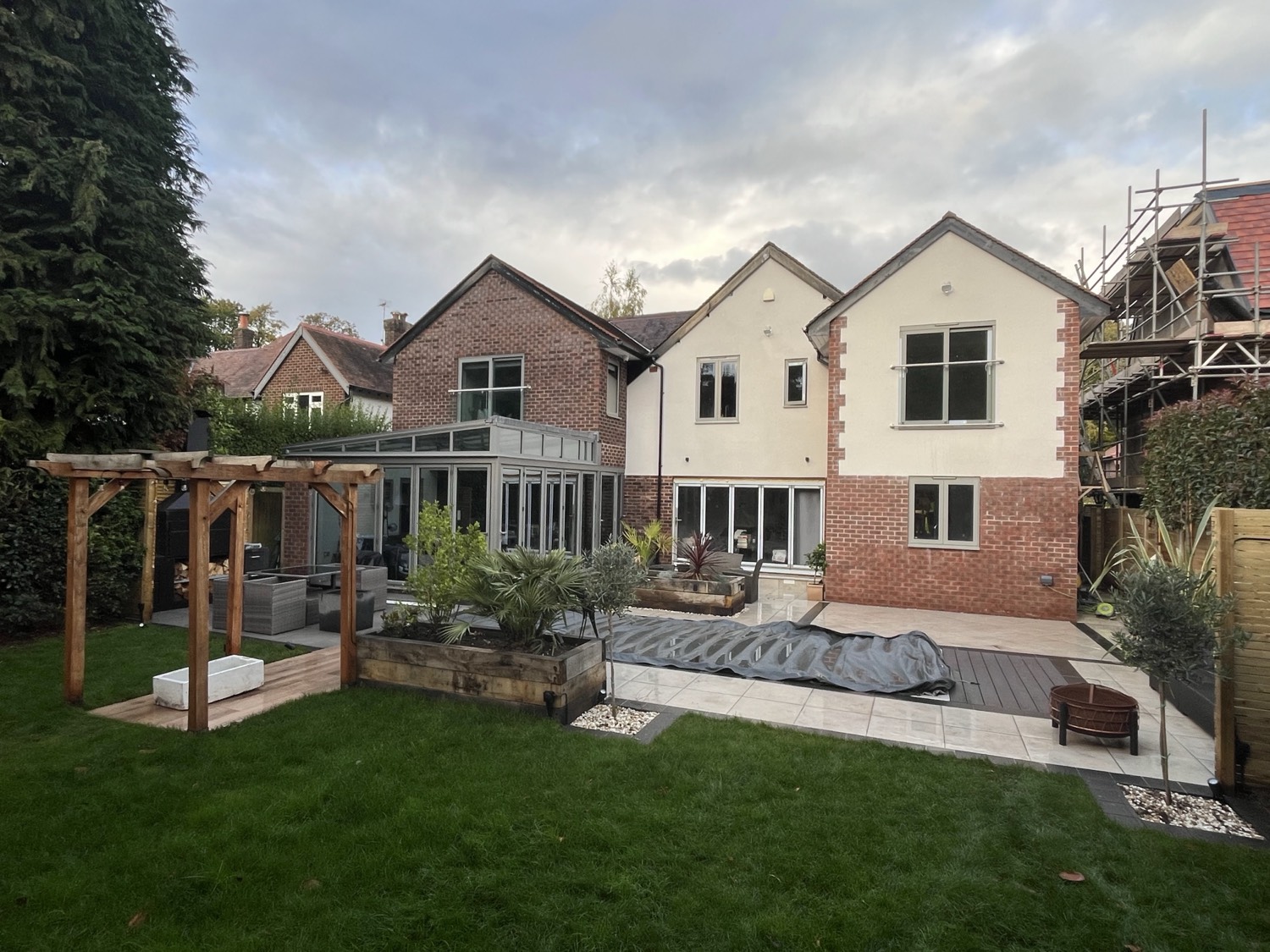 A contemporary rear garden with a raised timber pergola over a lounge area, sleeper-edged planters, artificial lawn and a porcelain terrace off the house.